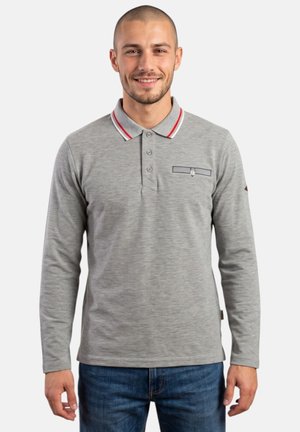 Homme souriant portant un polo gris à manches longues avec un col rayé rouge et blanc et un jean bleu, debout sur fond blanc.