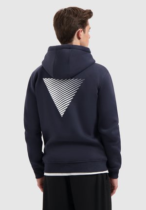 Junger Mann, der von hinten schaut, trägt einen navyblauen Kapuzenpulli mit einem weißen gestreiften Dreieckmuster auf dem Rücken und schwarze Hose.