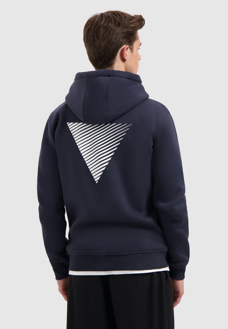 Jonge man die van zich af kijkt, gekleed in een marineblauwe hoodie met een wit gestreepte driehoekfunctie op de rug en zwarte broek.