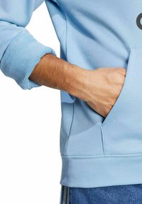 Hoodie di cotone azzurro chiaro con tasca frontale, polsini e bordo a costine. Il tessuto è liscio con una sottile texture. Mano parzialmente visibile nella tasca.