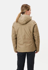 Khaki pufferjack met capuchon, met een gladde textuur, gestikte details en een iets verlengde achterzoom. Donkere broek zichtbaar eronder.