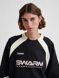 Young woman with short brown hair wearing black and white sweatshirt with "SWARM INTERNATIONAL" and "hummel sportswear" logos on a plain background.