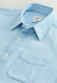 Chemise légère bleu ciel à boutons, avec col pointu et poche poitrine, en tissu semblable au denim, taille moyenne avec étiquette de marque visible.