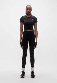 Γυναίκα που στέκεται φορώντας μαύρο αθλητικό crop top, κολάν, γάντια χωρίς δάχτυλα, λευκές κάλτσες και μαύρα αθλητικά παπούτσια σε λευκό φόντο.