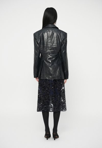 Czarna skórzana marynarka z usztywnionymi ramionami i szwem na plecach, zestawiona z granatową spódnicą midi zdobioną cekinami, uzupełniona czarnymi rajstopami.