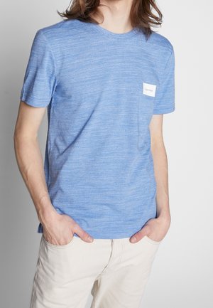 Person iført en lyseblå kortærmet skjorte med et lille hvidt Calvin Klein-logo på brystet og lyse beige bukser med hænderne i lommerne.