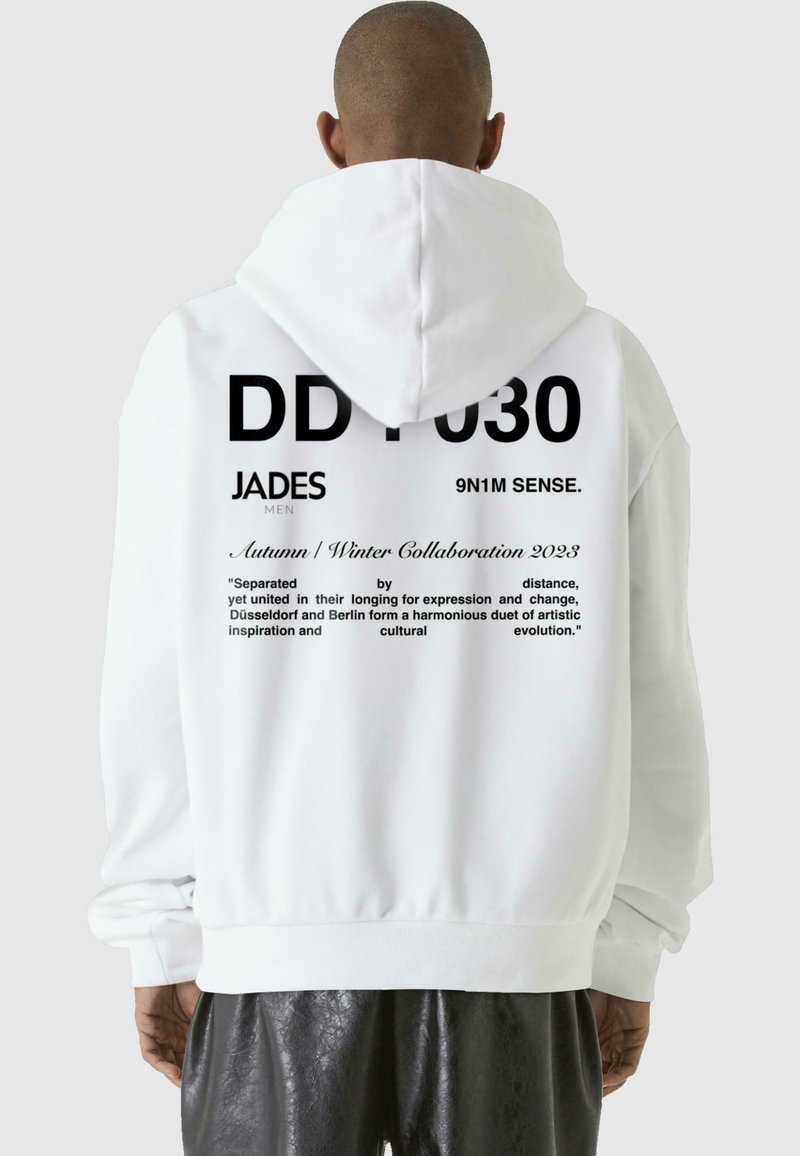 Valge kapuutsiga sviiter ees koti ja musta tekstiga seljal, mis sisaldab disainielemente ja logosid. Tekst sisaldab tsitaati ja kollektsiooni teavet.