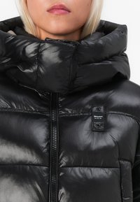 Schwarze Pufferjacke mit glänzender, gesteppten Textur, hohem Kragen und vollem Reißverschluss. Mit Logo-Patch auf der linken Seite nahe dem Hals.