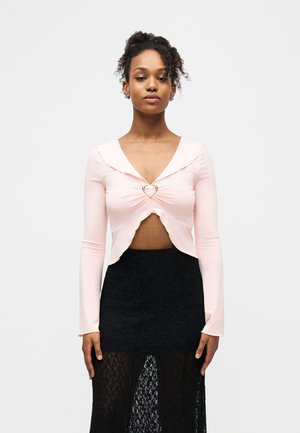 Γυναίκα με κατσαρά μαλλιά που φοράει ανοιχτό ροζ κοντομάνικο τοπ με κούμπωμα σε σχήμα καρδιάς και μαύρη φούστα από δαντέλα, στέκεται μπροστά σε μια λευκή επιφάνεια.