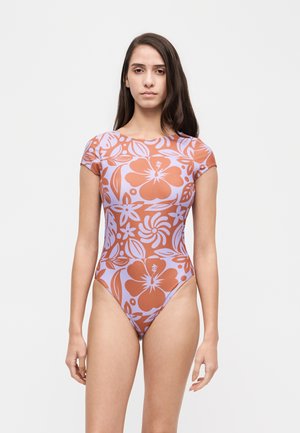 Une femme porte un maillot de bain à manches courtes, à imprimé floral lilas et corail, sur un fond clair uni.