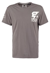 Šedé tričko s krátkým rukávem s bílým logem "G RAW" a hvězdou na levé horní části hrudi a malým bílým textem na pravém rukávu.
