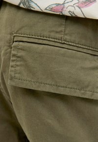 Pantalons vert olive avec une poche arrière à rabat. Le tissu a une texture lisse, avec des coutures visibles le long des bords et de la poche.