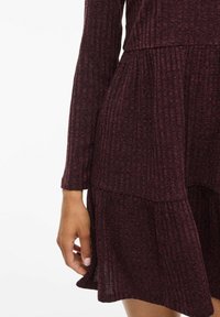 Robe tricotée bordeaux à manches longues, ornée d'un design à étages et d'un tissu texturé. Les détails plissés à la taille mettent en valeur la silhouette.