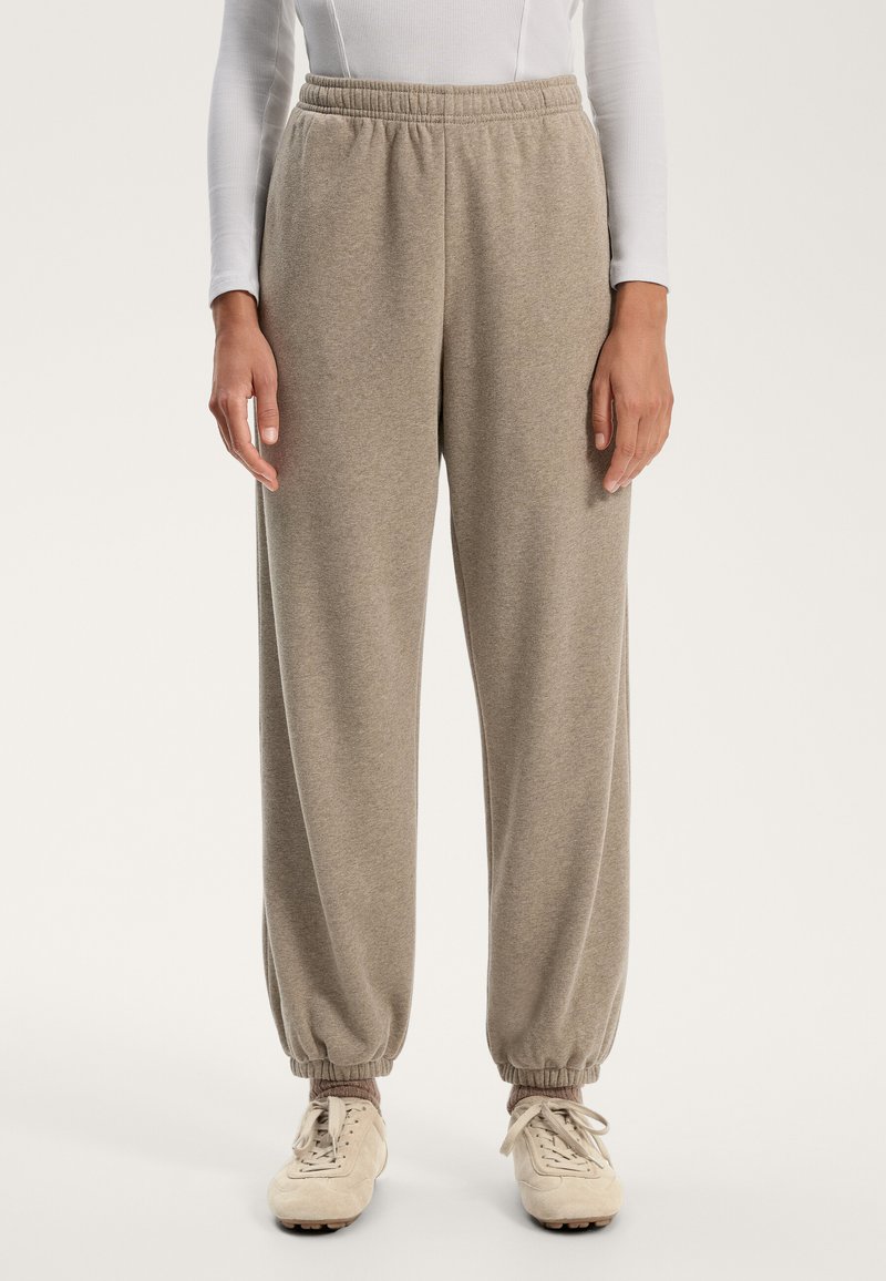 Vaaleanruskeat collegehousut pehmeästä materiaalista, joissa on joustava vyötärö ja kuminauha lahkeissa, yhdistettynä vaaleankermanvärisiin lenkkareihin.