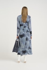 Vestido longo azul com padrão abstrato, mangas longas e costas franzidas, combinado com uma mala cinza e botas brancas até a coxa.