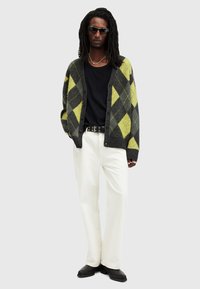 Cardigan con motivo argyle in grigio scuro e giallo neon, indossato sopra una maglietta nera. Abbinato a pantaloni bianchi e stivali neri.