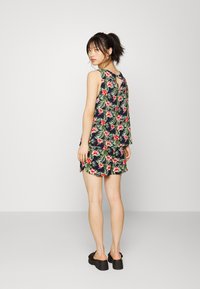 Macacão de estampa floral em azul-marinho escuro com folhas verdes e flores vermelhas; apresenta um decote em forma de lágrima nas costas, design sem mangas e bainha assimétrica.