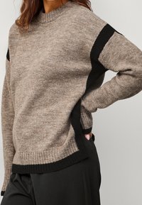 Pull en tricot beige avec des accents noirs, doté d'un col rond, d'un ourlet côtelé et de fentes latérales pour un confort accru. Texture douce.