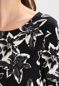 Haut noir avec un motif floral en blanc et gris. Encolure ronde, texture lisse et manches courtes.