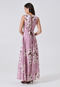 Frau, die ein ärmelloses, lavendelfarbenes Maxikleid mit floralem Muster und Rückbanddetail trägt, steht vor einem schlichten, hellen Hintergrund.