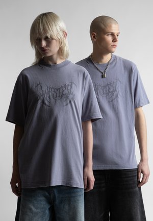 Dwie osoby noszące przyduże szare koszulki z nadrukiem stoją obok siebie na neutralnym tle, jedna o blond włosach, druga z ogoloną głową.