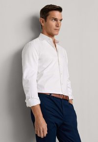 Mann trägt ein weißes langärmliges Hemd mit aufgekrempelten Ärmeln, marineblaue Hose und braunen Gürtel, steht vor einer hellen Wand.