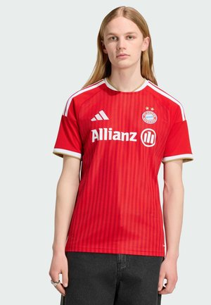 Joven con cabello largo y rubio lleva una camiseta roja del Bayern Munich con bordes blancos y dorados y pantalones negros sobre un fondo liso.