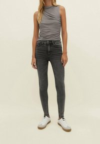 Blusa gris sin mangas con detalle fruncido, combinada con jeans skinny negros de cintura alta. Los zapatos son blancos con acentos grises. Fondo simple.