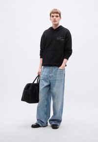 Μαύρο φούτερ με κουκούλα και μπροστινή τσέπη με λογότυπο, συνδυασμένο με φαρδιά τζιν παντελόνια ανοιχτού μπλε χρώματος. Το μοντέλο κρατά μια μαύρη τσάντα και φοράει μαύρα παπούτσια.