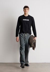 Camisa de mangas longas preta com o logótipo "Calvin Klein", jeans cinzentos claros e sapatos escuros. Asegurando um suéter castanho xadrez. Fundo neutro.