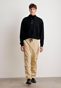 Svart langermet polo skjorte med tre knapper, kombinert med beige cargo-bukser med lommer og strikk. Svarte sko fullfører antrekket.