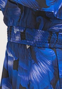 Robe fleurie bleue avec une texture froissée, taille smockée et une petite étiquette métallique. Le design intègre différentes nuances de bleu.