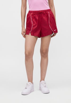 Mujer con pantalones cortos deportivos rojos brillantes con ribete blanco, zapatillas blancas Nike y una camiseta sin mangas rosa, de pie frente a un fondo liso.