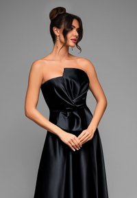 Musta satiinikangas strapless-mekko, jossa on kierretty muotoilu rinnassa, virtaava hame ja elegantti kiilto. Malli seisoi kädet ristissä.