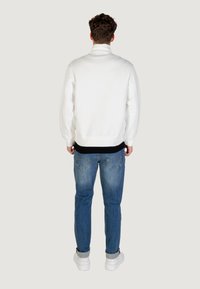 Sudadera blanca con cuello alto y puños acanalados, combinada con jeans de color azul claro y zapatillas blancas, vista desde la parte de atrás.