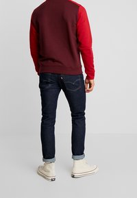Uomo che indossa jeans blu scuri con risvolto, una maglietta a maniche lunghe bordeaux e rossa, e sneakers bianche alte, in piedi su uno sfondo semplice.