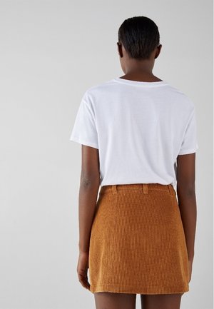 Persona de espaldas vistiendo una camiseta blanca de manga corta metida en una falda marrón de pana de talle alto, sobre un fondo liso.