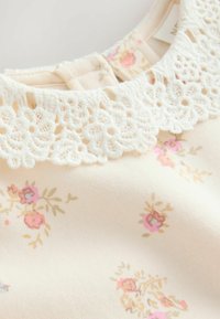 Indumento color crema con un colletto di pizzo bianco texturizzato. Il tessuto presenta un motivo floreale con accenti rosa e beige.