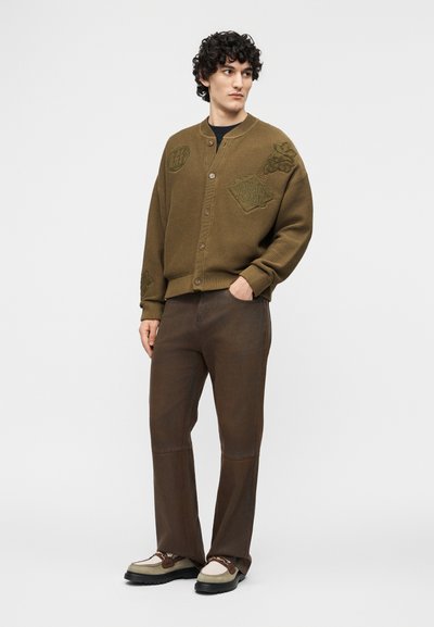 Άνδρας με σγουρά μαλλιά, που φοράει ένα καφέ πλεκτό ζακέτα με κουμπιά και μπαλώματα, σκούρο καφέ παντελόνι και μπεζ και καφέ αθλητικά παπούτσια, στέκεται με το ένα χέρι στην τσέπη.