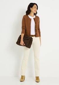 Cardigan marrón acolchado con botones delanteros, camiseta blanca, pantalones crema y botines beige. Sosteniendo un bolso de ante marrón oscuro.