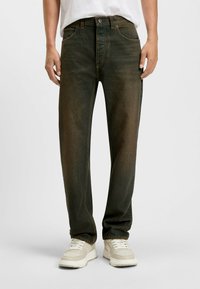 Vaqueros de mezclilla verde con un corte recto, que cuentan con cinco bolsillos, cierre de botón y detalles de costura en contraste. Combinados con zapatillas claras.