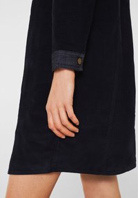 Femme portant une robe en velours côtelé bleu foncé avec des poignets roulés et un bouton en laiton, montrant la main et une partie des jambes nues.