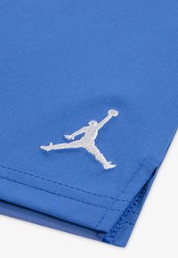 Modrý materiál má vyšívané bílé logo Jumpman. Textilie je hladká s jemným leskem, vykazuje pečlivé prošívání a čisté okraje.