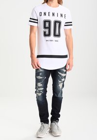 Bílý tričko s krátkým rukávem a černými akcenty, s číslem "90" na přední straně, v kombinaci s roztrhanými tmavými džíny a světlými teniskami.