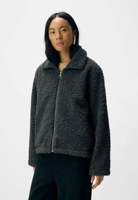 Veste en polaire de couleur gris foncé, avec une fermeture éclair à l'avant, un col haut et une surface texturée. Coupe loose avec des manches longues et une poche latérale.