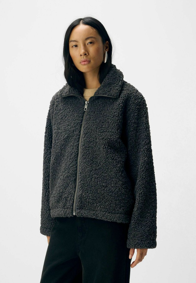 Veste en polaire de couleur gris foncé, avec une fermeture éclair à l'avant, un col haut et une surface texturée. Coupe loose avec des manches longues et une poche latérale.