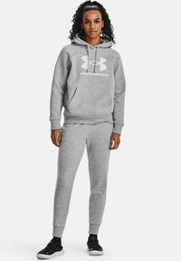 Szary bawełniany hoodie z dużym białym logo Under Armour, zestawiony z pasującymi szarymi dresami i czarnymi butami sportowymi.