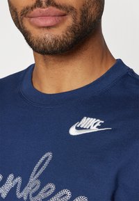 Sudadera azul marino con cuello redondo acanalado, que presenta un logo de Nike bordado en blanco y un diseño de texto en blanco texturizado en la parte delantera.
