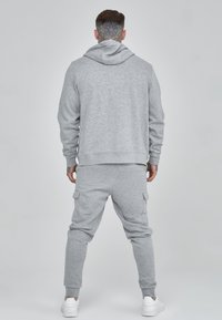 Grauer Sweatshirt mit Kapuze, kombiniert mit passenden Cargo-Hosen. Weicher Stoff, slim-fit, elastische Bündchen und Seitentaschen. Weiße Sneakers sichtbar.