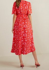 Robe midi florale rouge avec manches courtes, taille froncée et jupe fluide. Présente des fleurs roses et blanches sur un tissu léger.